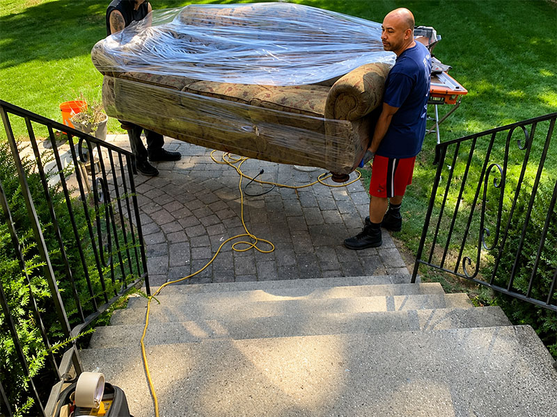 Brother Movers crew carefully carrying a shrink-wrapped sofa down outdoor stairs during a residential move in Lawrence, MA
